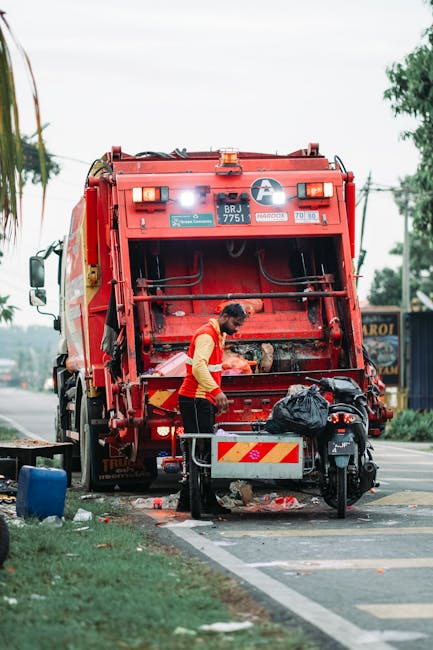 A waste collection worker dressed in a high-visibility yellow and red vest is operating a large red waste disposal vehicle parked on the side of a street. The worker is leaning forward, handling debris or waste within the open rear compartment of the truck. The vehicle's back section is raised, revealing internal compartments and mechanical components, with a small warning light on top. In front of the truck, a black motorcycle with a plastic bag secured on the seat is positioned close to a rectangular metal barrier or bin. Scattered waste or debris is visible on the pavement beneath the worker, along with a blue container nearby. The setting appears to be an outdoor urban area during daytime, with a paved road, some greenery, and distant trees visible under an overcast sky, illustrating a scene of on-site rubbish collection as part of an independent waste removal service, with Waste Clearance Ealing potentially involved in the process.