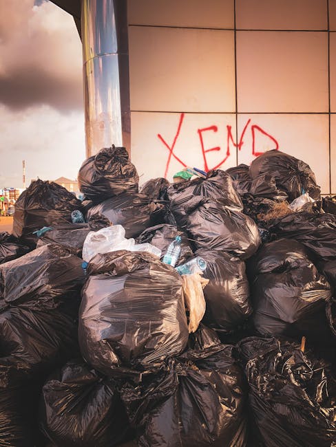 A large pile of black rubbish bags filled with waste material stacked against a modern building with white tiled exterior walls. The bags appear to contain household refuse and are placed on a paved surface, likely a sidewalk or driveway. Several transparent plastic water bottles are visible protruding from some of the bags, with crumpled plastic wrappers and other small debris scattered around the base. In the background, part of the cityscape and cloudy sky are visible, with the scene illuminated by natural, possibly late afternoon or early evening, light. The bags are unarranged, with some leaning against the wall and others piled haphazardly, indicating an off-site clearance process. The setting suggests an activity related to independent waste disposal, possibly by a private rubbish removal service such as Waste Clearance Ealing, operating in the vicinity of an urban commercial or residential area.