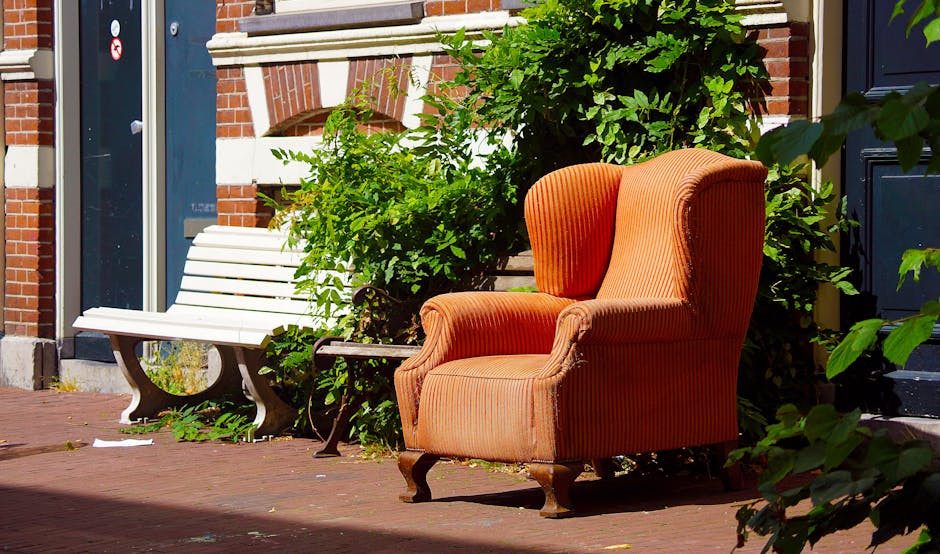 The image shows an orange upholstered armchair with a rounded back and wooden cabriole legs positioned on a brick pavement exterior of a building, adjacent to a white wooden bench with curved black metal armrests. Behind the armchair, dense green foliage and leafy bushes partially obscure the backdrop of a brick wall with decorative brickwork and white stone accents. The scene is illuminated by natural daylight, creating subtle shadows on the ground. The presence of a private armchair outdoors next to a bench suggests an informal area potentially used for seating or relaxation, contrasted with typical rubbish removal scenes. The setting appears to be in an urban or residential environment with the armchair and bench located on a street or courtyard, which Waste Clearance Ealing may assist with in terms of alternative waste disposal or on-site clearance of unwanted furniture and garden waste, reflecting the company's rubbish collection services.