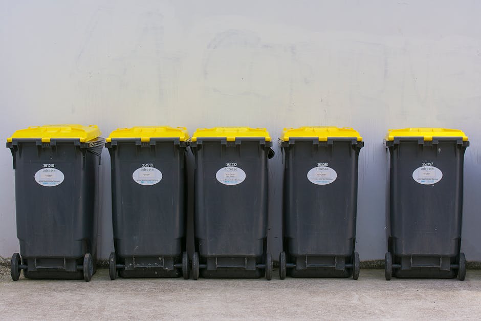 Five large wheeled rubbish bins with black bodies and bright yellow lids are lined up in a row on a paved surface against a plain, light-colored wall. Each bin has a white oval label with printed identification numbers and details, positioned in the upper middle section of the bin's front face. The bins are positioned close together, with the wheels visible at the bottom, indicating they are for waste collection or disposal, likely used for private refuse management. The scene is evenly lit, capturing the clean, modern appearance of the bins and the neutral environment surrounding them, reflecting typical equipment used by waste clearance services such as Waste Clearance Ealing in their alternative waste handling or on-site rubbish removal activities.