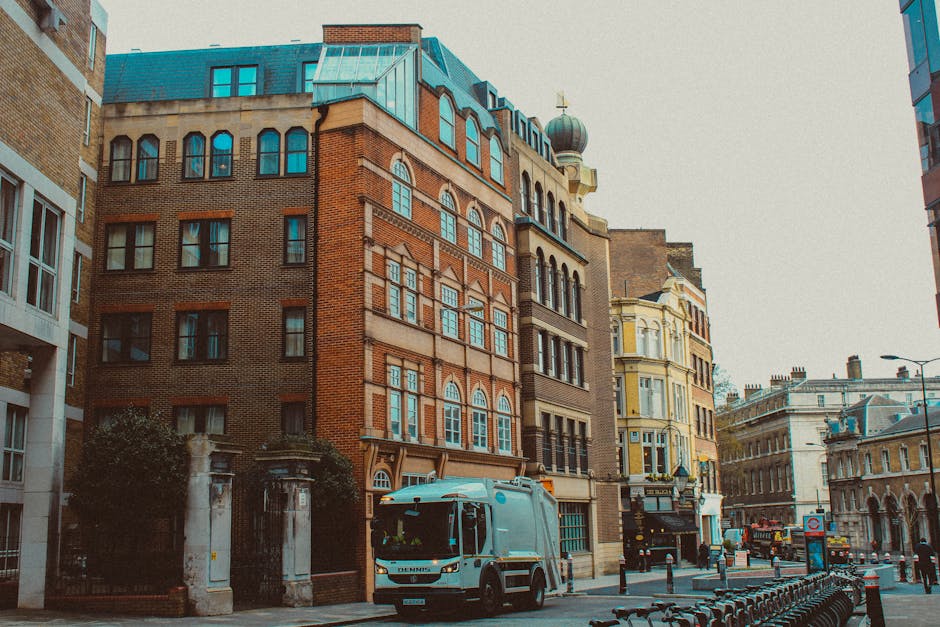 A street scene featuring a grey waste collection truck operated by Waste Clearance Ealing parked on the pavement in front of a set of historic multi-storey buildings with ornate architectural details, large sash windows, and a dome structure on the roof. The truck's side panel and front grille are visible, with the vehicle positioned close to the curb. Surrounding the scene are other buildings with varied facades in brick and painted finishes, along with street furniture such as bike racks, bollards, and a street lamp. The environment appears urban with clear weather and natural daylight illuminating the scene, suggesting a typical city environment suitable for private rubbish collection and on-site waste clearance services conducted by waste management specialists like Waste Clearance Ealing.
