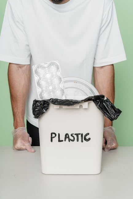 A person wearing a white t-shirt and transparent gloves is holding a white recycling bin labeled 'PLASTIC' with black marker, which is partially covered with a black rubbish bag. Inside the bin, there is a clear plastic egg carton with six compartments and a white plastic container or lid, both visible above the bin's rim. The background is a plain, light green wall. The scene emphasizes waste segregation and the collection of plastic materials, typical of private or alternative rubbish removal services like those offered by Waste Clearance Ealing, focusing on environmentally conscious waste handling and on-site collection options.