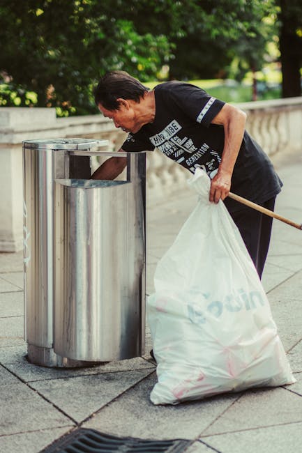 A person disposing of brown paper bags into a white bin for rubbish collection, with additional paper bags in hand. The bin has a partially visible label indicating it is used for waste disposal. The scene is set indoors, with a wooden surface nearby and a dark interior background. The paper bags vary slightly in size and are crumpled or folded, made of plain, brown kraft paper with a slightly rough texture. The person's arm is visible, wearing a short-sleeved shirt, and the hand is grasping the paper bags, ready to place them inside the bin. The lighting is soft and natural, highlighting the different textures of the paper and the smooth surface of the bin. This image relates to waste management activities, such as household rubbish removal, illustrating the process of segregating waste for disposal or private collection services like those offered by Waste Clearance Ealing, which provides alternative waste handling options beyond municipal collection.
