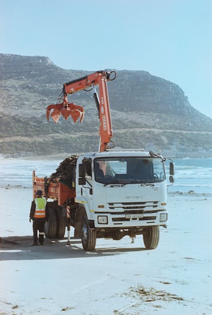 A white waste collection truck equipped with an orange hydraulic arm is parked on a sandy beach, with a worker in high-visibility clothing standing beside it. The hydraulic arm extends upward and outward, holding a grapple attachment filled with dark, loose debris, likely soil or construction waste. The truck's cab features side mirrors and a front grille, with the body showing signs of use. The background reveals a coastal landscape with a rocky cliff face and grassy slopes, under a clear blue sky. The scene suggests an on-site waste clearance operation, possibly related to environmental or construction work, performed by a waste management service such as Waste Clearance Ealing that offers alternative private waste disposal options outside typical urban settings, although the location appears to be remote from urban centre infrastructure.