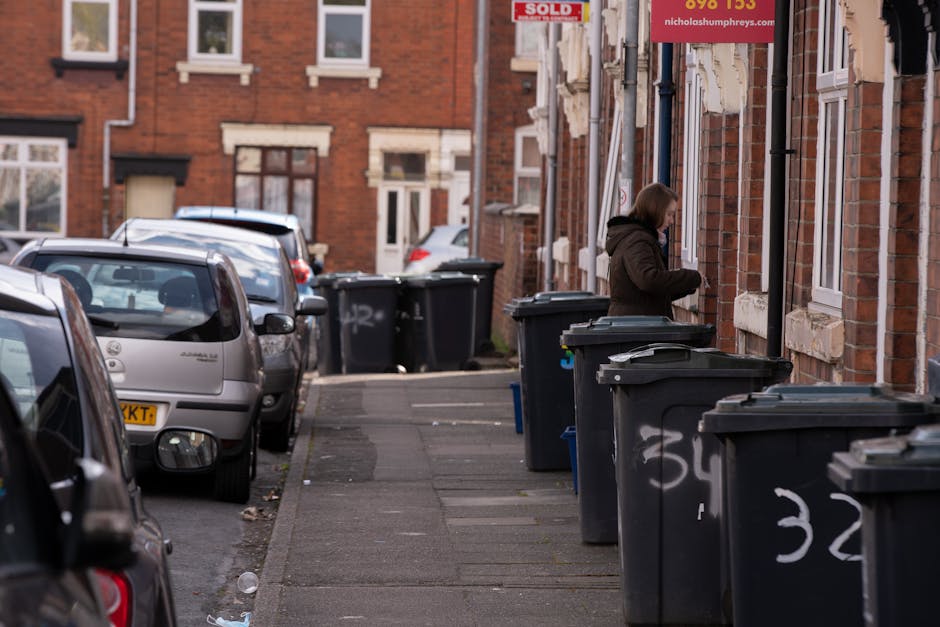 A woman standing on a narrow residential pavement beside a row of large black wheelie bins with no visible markings, positioned against the red-brick exterior of terraced houses. The bins have dark grey lids, with some lids slightly open, and are situated close to the wall, in front of white-framed sash windows. The woman appears to be using her phone or a small device, with her head slightly bent down. To the left, parked cars of various makes and models, including a silver hatchback and a dark vehicle, are lined up along the street, extending towards the background. The street is paved with grey concrete slabs, and the surrounding environment features a typical urban setting with brick buildings, some with signs, such as a partly visible 'SOLD' sign and a smaller red sign with contact details. The overall scene suggests typical bin storage outside residential properties, consistent with private waste collection practices, with an emphasis on rubbish removal seen through the presence of multiple bins awaiting collection or disposal within a local waste management context.